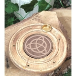 Porte-encens bois rond triquetra, posé sur tranche de bois brut avec feuillage vert en fond, ambiance calme et protection symbolique.