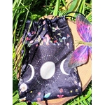 Pochon de rangement en velours noir, symbole triple lune blanc (croissant, pleine lune, croissant) sur l’avant, cordons de serrage en haut, posé sur planche de bois, entouré de feuillage vert, lumière naturelle, ambiance mystique et naturelle.