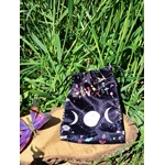 Pochon de rangement en velours noir motif galaxie, symbole triple lune blanc sur l’avant, présenté sur rondin de bois avec feuillage vert en arrière-plan, papillon décoratif violet sur le côté, lumière naturelle, ambiance nature et mystique.