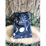 Pochon de rangement en velours noir motif ciel étoilé, symbole triple lune blanc sur l’avant, présenté sur rondin de bois devant une écorce d’arbre, avec feuillage vert en décor, lumière naturelle, ambiance nature et ésotérique.