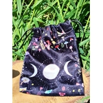 Pochon de rangement en velours noir motif galaxie, symbole triple lune blanc sur l’avant, cordon de serrage visible, posé sur planche de bois avec grandes feuilles vertes en arrière-plan, lumière naturelle, ambiance ésotérique et nature