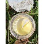 Vue du dessus d’une bougie en cire de soja couleur crème, mèche en coton au centre, dans un pot en verre, posée dans l’herbe avec amas de quartz en arrière-plan, ambiance nature, clarté, purification, apaisement, recentrage, douceur.