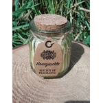 Bougie parfumée au chèvrefeuille en cire de soja, en pot en verre avec couvercle en liège, posée sur tissu beige avec feuillage vert en arrière-plan, ambiance naturelle, douceur, réconfort, apaisement, harmonie, cocooning.