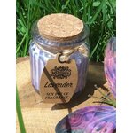 Bougie lavande en pot de verre violet strié avec couvercle en liège et étiquette kraft, posée sur planche en bois en extérieur, herbes en fond, ambiance douce et relaxante.