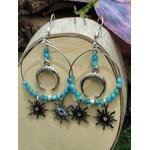 Boucles d’oreilles Atlante en apatite bleue, création Magic Gaïa, anneaux argentés avec symbole lune et charms soleil, présentées sur support en bois clair en intérieur. Apaisement, communication, clarté intérieure, confiance douce