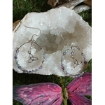 Boucles d’oreilles Hécate en fluorite facettée, création Magic Gaïa, anneaux perlés dégradés et triquetra argentée, posées sur quartz blanc avec ailes de papillon roses en décor. Clarté, apaisement, intuition