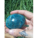 Pierre roulée d’apatite bleue poli bleu lagon marbré, tenue entre les doigts devant des herbes vertes, pierre naturelle liée à l’authenticité, au recentrage et à la fluidité d’expression.