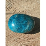 Pierre roulée d’apatite bleue aux nuances bleu lagon marbrées, posée sur support beige texturé avec ombre portée, pierre naturelle associée au discernement, au calme du mental et au dynamisme doux.