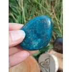 Galet d’apatite bleue poli tenu entre les doigts, avec herbes vertes et décor ésotérique en arrière-plan, pierre naturelle liée au recentrage, à l’authenticité et à la sensibilité apaisée.