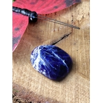 Pierre roulée de sodalite bleu nuit veinée de blanc sur bois naturel avec plume et décor sombre, galet poli évoquant la profondeur intérieure, la parole fluide et le calme du mental.