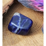 Pierre roulée de sodalite bleu nuit aux veines blanches, galet poli carré posé sur tranche de bois en décor naturel, pierre associée à la concentration paisible, à l’assurance et à la parole juste.