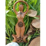 Statuette Gaïa en bois style wicca avec spirale et triquetra, mise en scène dans un décor feuillage avec galets et rondins; suggère ancrage, sérénité, harmonie, protection douce et connexion à la nature.