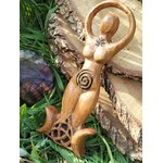 Statuette Gaïa en bois style wicca, spirale gravée et triquetra sur base triple lune, posée contre une écorce avec feuillage et pierre violette; évoque enracinement, protection, équilibre, intuition et lien à la Terre.