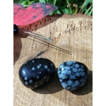 Deux pierres roulées d’obsidienne neige noir moucheté blanc sur tranche de bois en extérieur avec herbes vertes et pierre violette, pierres naturelles liées à la maîtrise émotionnelle, à la stabilité et à la tranquillité.