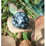 Pierre roulée d’obsidienne neige noir profond à motifs blancs floconnés, posée sur tranche de bois en extérieur avec herbes vertes, pierre naturelle associée à la sérénité, à la présence à soi et à l’ancrage.