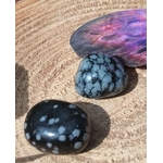 Deux pierres roulées d’obsidienne neige noir moucheté blanc posées sur une tranche de bois avec pierre violette en arrière-plan, pierres naturelles évoquant la stabilité intérieure, le retour au calme et l’ancrage doux.