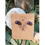 Boucles d’oreilles puces améthyste fixées sur une carte kraft avec motif attrape-rêves, posée devant une coquille blanche, décor d’herbes vertes en arrière-plan; évoque calme, douceur, protection (tradition).