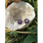 Boucles d’oreilles puces améthyste en duo sur pierre blanche irrégulière, brins verts qui traversent le cadre, photo en extérieur; évoque apaisement, harmonie, douceur.