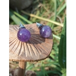 Boucles d’oreilles puces améthyste violette sur tranche de bois brut, soleil rasant et ombres marquées, arrière-plan végétal; associée à calme intérieur, équilibre émotionnel, intuition.