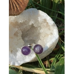 Boucles d’oreilles puces améthyste posées côte à côte sur un amas de quartz, brins d’herbe et teintes naturelles en fond; associée à équilibre émotionnel, paix intérieure, protection (tradition).