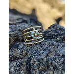 Bague dorée acier inoxydable style spirale multi anneaux, grappe de cristaux bleus au centre, mise en scène sur rocher granitique, fond mer et plage en flou artistique, photo en Bretagne.