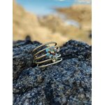 Bague dorée multi-anneaux torsadés acier inoxydable avec cristaux bleus, posée sur rochers sombres en bord de mer, plage et eau floutées en arrière-plan, photo prise en Bretagne.