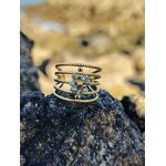 Bague dorée à plusieurs rangs texturés en acier inoxydable, ornée de petits cristaux bleus irisés, mise en scène sur rocher noir en bord de mer, fond sable flou, photo en Bretagne.