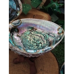 Coquille d’ormeau nacrée aux reflets bleu-vert/violet, posée sur bois avec feuillage en arrière-plan; support encens/fumigation, évoque purification, protection et calme