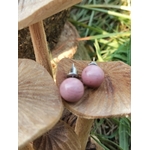 Boucles d’oreilles puces rhodonite rose, tiges acier inoxydable, posées sur support en bois avec feuillage en arrière-plan; douceur, apaisement émotionnel.