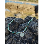 Collier en acier inoxydable doré avec cristaux verts et petites perles, pendentif rond doré au centre, photographié en Bretagne posé sur des rochers en bord de mer; évoque fraîcheur, harmonie et élégance naturelle.
