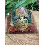 Pyramide orgonite 7 chakras avec fleur de vie et spirale de cuivre, posée sur une tranche de bois avec herbe en arrière-plan ; évoque harmonie, équilibre, ancrage.