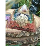 Pyramide orgonite 7 chakras avec fleur de vie et spirale de cuivre, posée sur une tranche de bois en extérieur (herbe, pierre au premier plan) ; évoque harmonie, ancrage, protection.