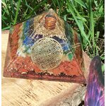 Pyramide orgonite 7 chakras avec fleur de vie et spirale de cuivre, posée sur tranche de bois en extérieur (herbe, pierre violette) ; évoque équilibre, ancrage, protection.