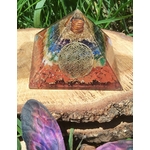 Pyramide orgonite 7 chakras avec fleur de vie et spirale de cuivre, posée sur une tranche de bois en extérieur (herbe, pierre violette au premier plan) ; évoque harmonie, protection, ancrage.