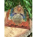 Pyramide orgonite 7 chakras avec fleur de vie et spirale de cuivre, posée sur tranche de bois en extérieur (herbe en arrière-plan) ; évoque équilibre, harmonisation, protection