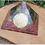Pyramide orgonite 7 chakras avec fleur de vie (métal) et spirale de cuivre, posée sur tranche de bois en extérieur (fond végétal) ; évoque stabilité, recentrage, protection.