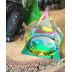 Pyramide en cristal irisé multicolore avec symbole yin et yang, posée sur une tranche de bois, avec une figurine noire et un décor vert flouté en arrière-plan ; aide à retrouver l’équilibre, calmer le mental et créer une ambiance harmonieuse.
