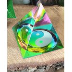 Pyramide en cristal irisé multicolore avec symbole yin et yang, posée sur une tranche de bois avec feuillage vert en arrière-plan ; invite à l’équilibre intérieur, la sérénité et l’harmonie des énergies.