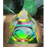 Pyramide en cristal irisé multicolore avec symbole yin et yang au centre, posée sur une tranche de bois, avec des feuilles vertes en arrière-plan ; apporte une ambiance d’équilibre, d’harmonie et d’apaisement.