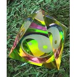 Pyramide en cristal irisé multicolore avec symbole yin et yang, posée sur un lit de feuillage vert ; apporte une sensation d’équilibre, d’harmonie et de calme intérieur.