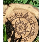 Dessous de plat rond en liège gravé d’un soleil et d’un croissant de lune, entourés de motifs végétaux; photo en extérieur dans l’herbe.