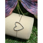 Bracelet chaîne fine en acier inoxydable, pendentif cœur ajouré; posé sur support blanc en extérieur, herbe en fond et papillon violet.