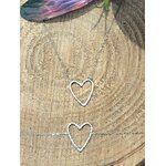 Deux bracelets chaîne fine en acier inoxydable avec pendentif cœur ajouré; posés sur rondin de bois, décor nature en extérieur.