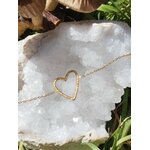 Bracelet chaîne fine en acier inoxydable doré, pendentif cœur ajouré; posé sur pierre blanche en extérieur, décor nature en arrière-plan.