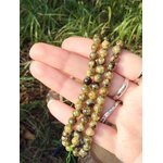 Fil de perles de péridot tenu en main sur fond d’herbe verte, nuances vert marbré et noir pour énergie lumineuse, renouveau et élan intérieur