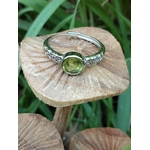 Bague réglable en cuivre blanc, péridot facetté vert rond, posée sur champignon en bois, fond de feuilles vertes ; évoque la joie, la clarté mentale, l’élan positif et l’équilibre émotionnel.