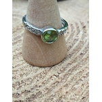 Bague réglable en cuivre blanc avec péridot facetté vert, posée sur un petit support en bois sur une rondelle de tronc ; apporte clarté, optimisme et apaisement.