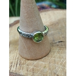 Bague réglable en cuivre blanc avec péridot facetté rond vert, portée sur un doigt, prise de vue rapprochée sur fond bois clair ; associée à l’optimisme, à la clarté d’esprit et à l’apaisement émotionnel.