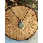 Pendentif garniérite vert-gris facetté sur cordon noir avec bélière argentée, posé au centre d’une tranche de bois aux cernes visibles, pierre naturelle associée à la douceur, à la stabilité et à l’harmonie émotionnelle.