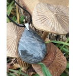 Pendentif goutte en pierre de lune noire polie sur cordon noir, photographié avec champignons décoratifs et feuillage en arrière-plan, bijou en pierre naturelle évoquant l’ancrage doux, le calme du cœur et la stabilité émotionnelle.
